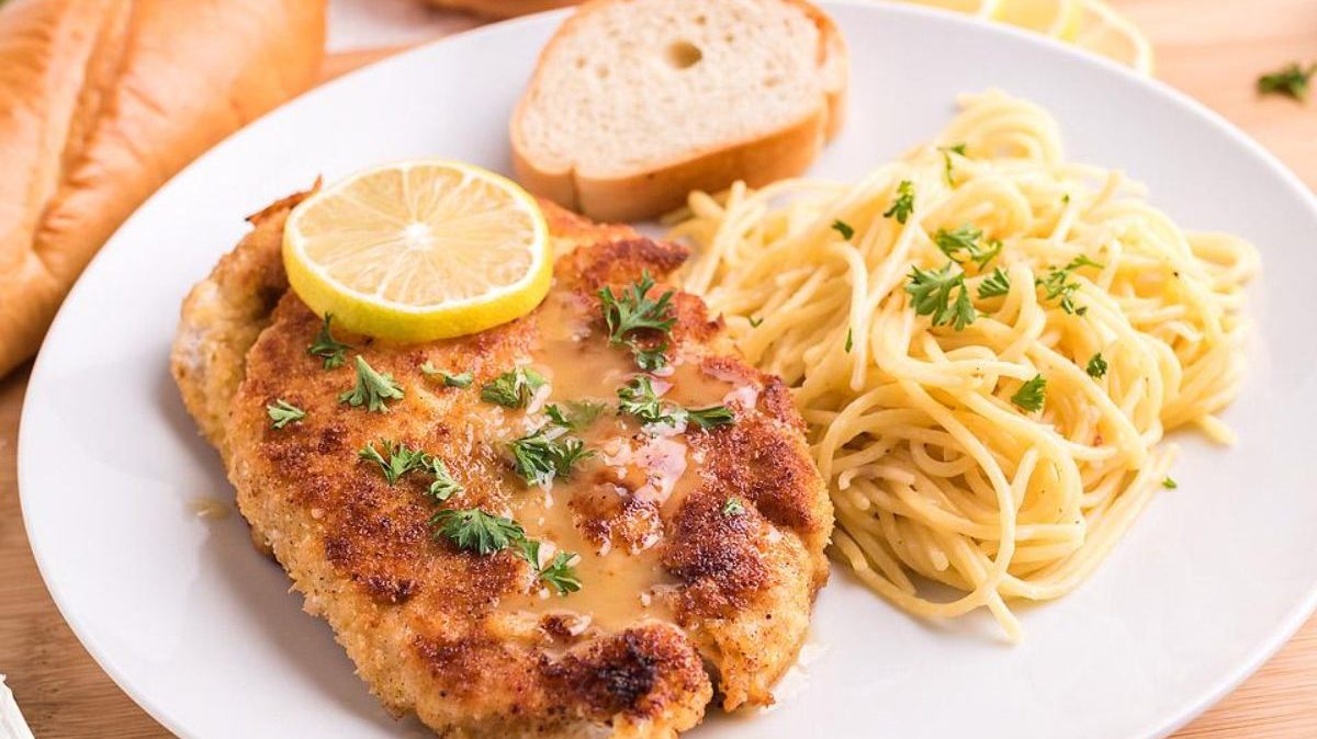 A plate with breaded chicken topped with lemon and sauce, served with a side of spaghetti garnished with parsley and a slice of bread in the background.