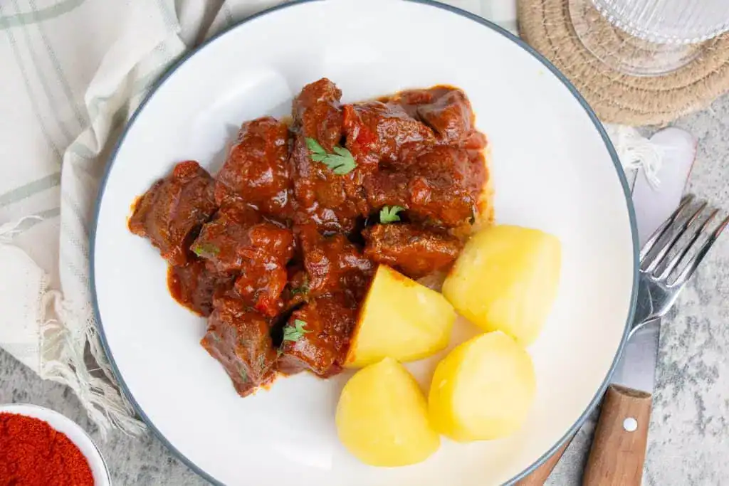 A white plate with beef stew in tomato sauce garnished with herbs and served with boiled potatoes. A fork and knife are placed beside the plate.