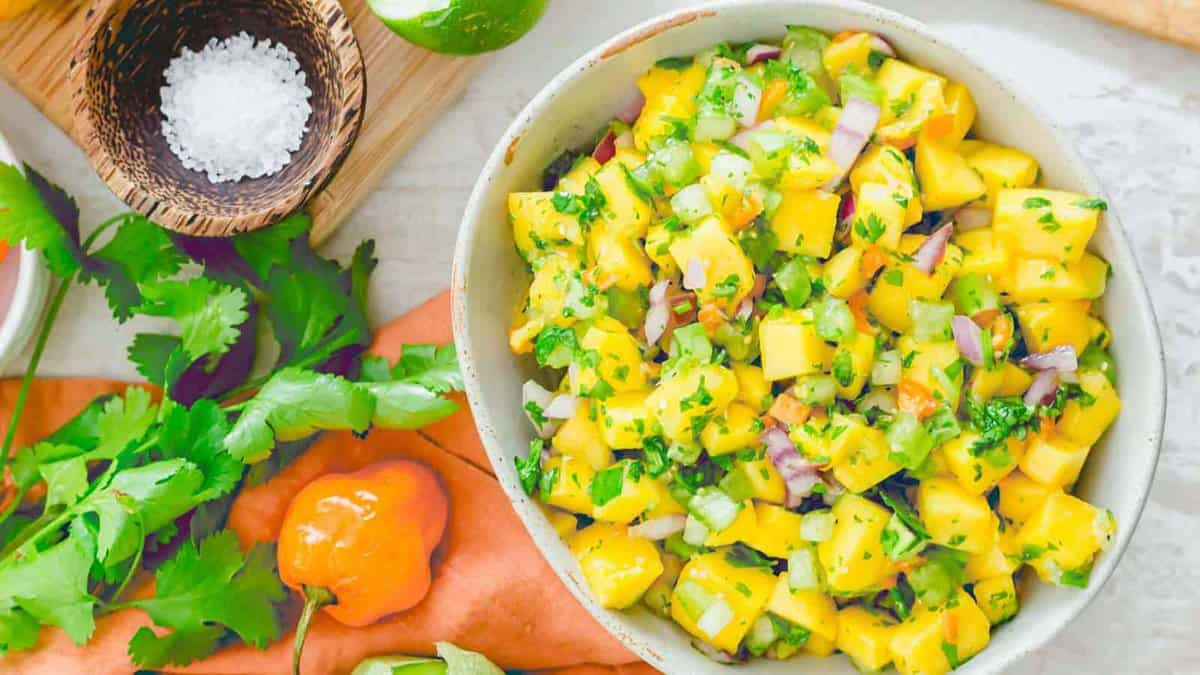 A bowl of mango salsa with diced mango, onion, cilantro, and peppers on a table, surrounded by fresh herbs, a habanero pepper, lime, and a bowl of salt.