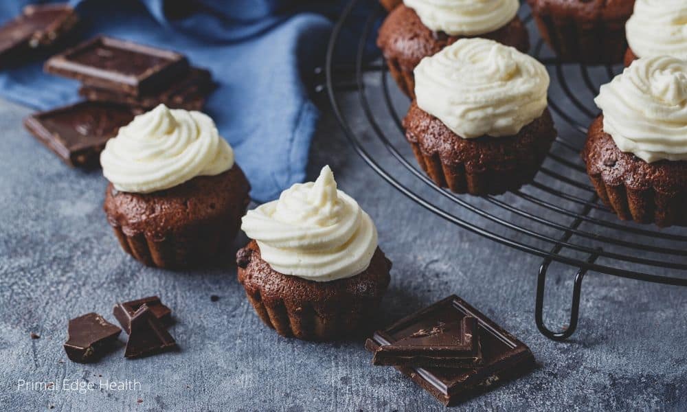 Chocolate cupcakes with white frosting sit on a cooling rack and a gray surface, with pieces of dark chocolate nearby.