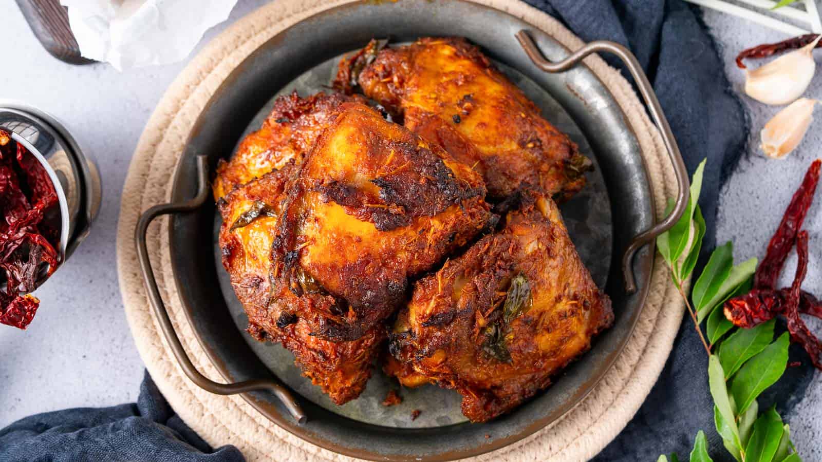 A metal plate with pieces of cooked, spiced chicken, surrounded by whole dried red chilies, garlic cloves, and green herbs on a light surface.
