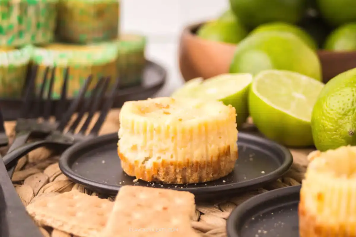 A mini cheesecake with a graham cracker crust is served on a black plate, surrounded by lime halves, graham crackers, and additional cheesecakes in the background.