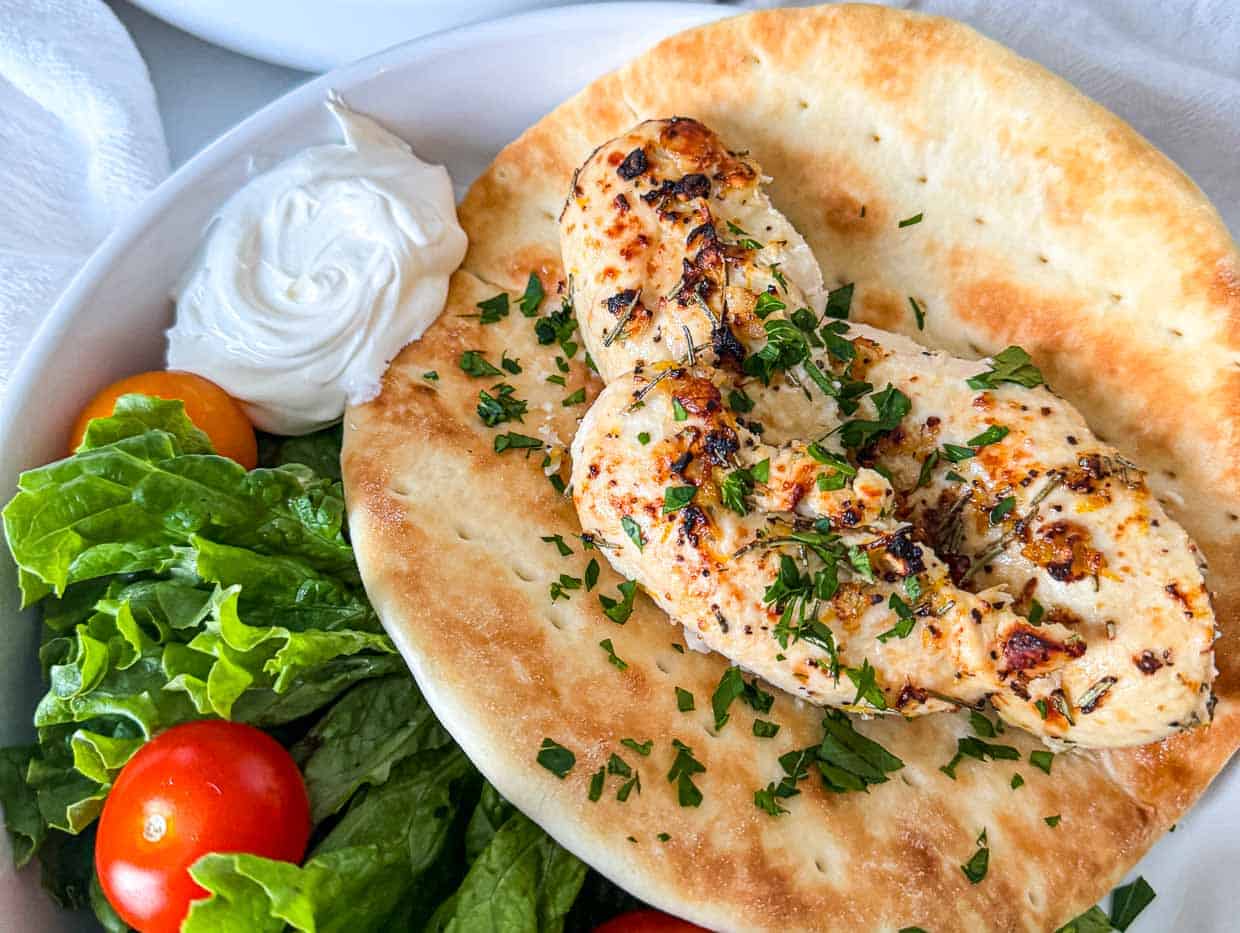 Grilled chicken breasts on flatbread, garnished with herbs, served with lettuce, cherry tomatoes, and a dollop of white sauce on a white plate.