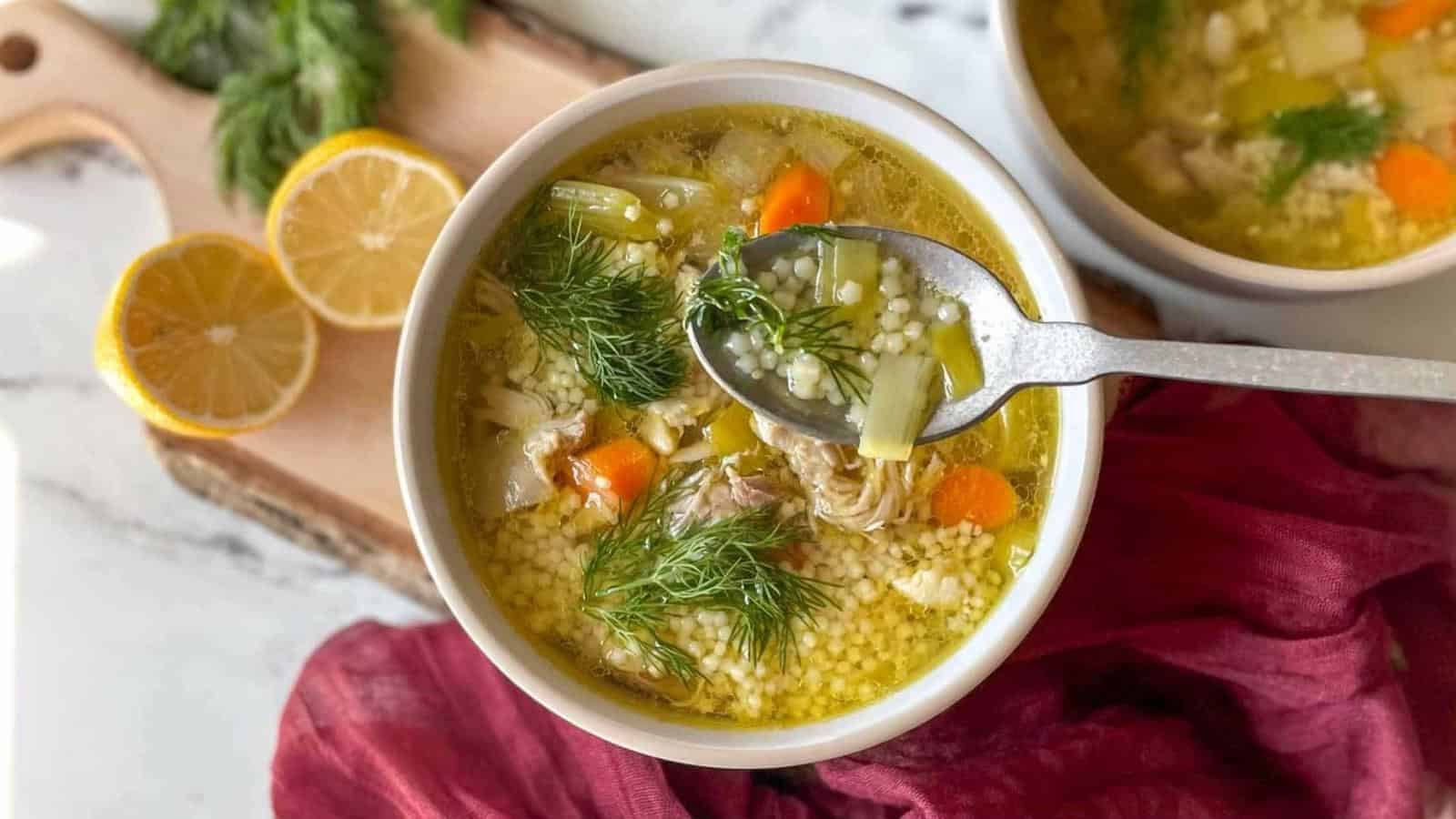 A bowl of chicken and vegetable soup with couscous, garnished with fresh dill. A spoon holds a portion above the bowl. Sliced lemon and herbs are on a wooden board nearby.
