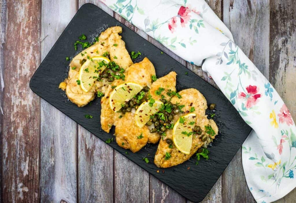 Three pieces of cooked chicken breast topped with lemon slices, capers, and chopped parsley, served on a black rectangular plate next to a floral napkin.