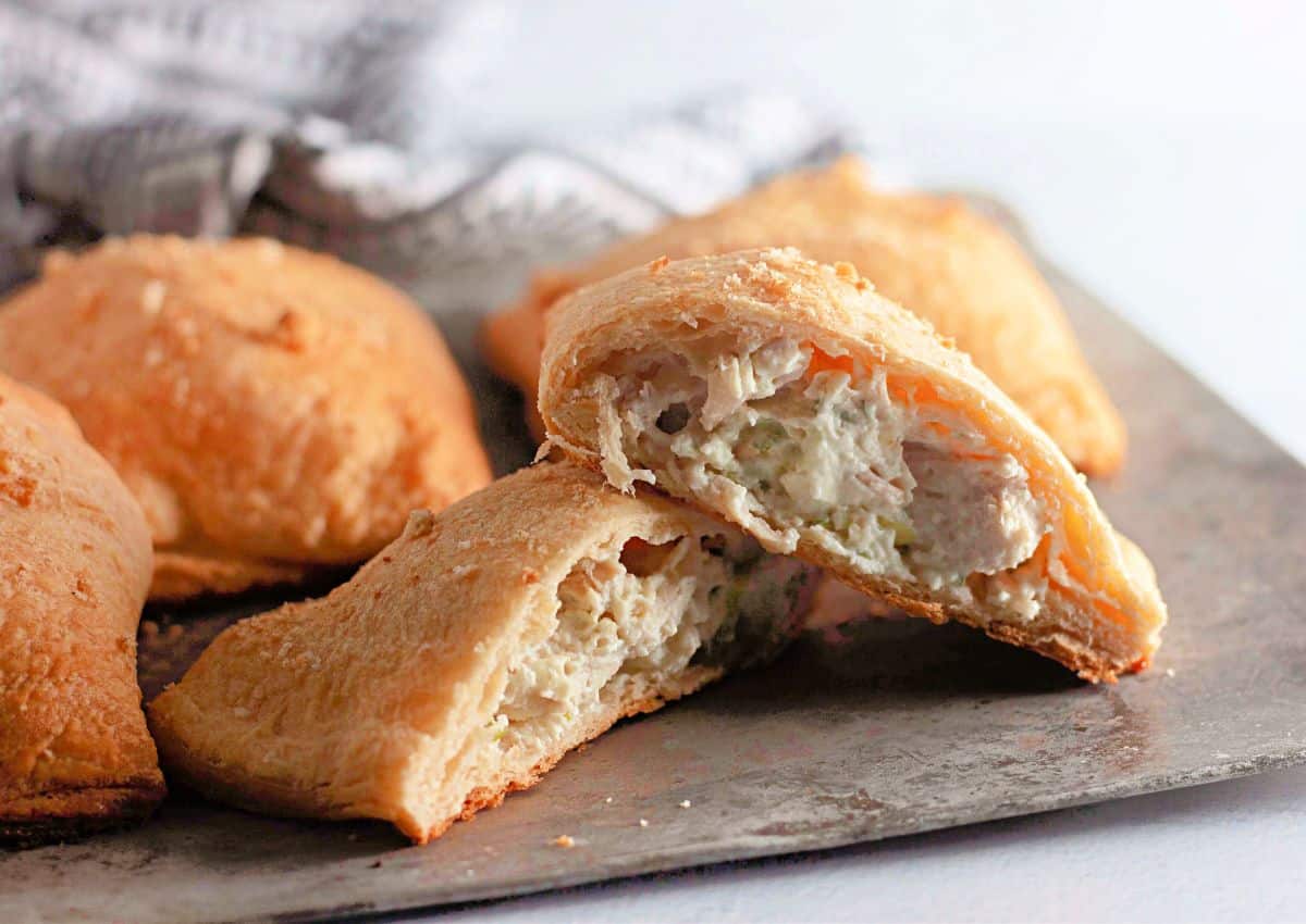 Golden-brown baked turnovers with a creamy chicken and vegetable filling, one cut open to show the inside, on a metal baking sheet.