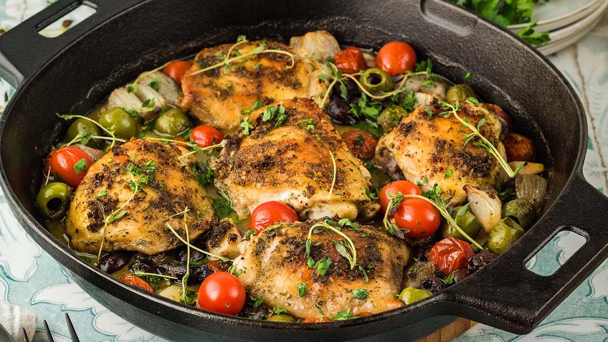 A cast iron skillet filled with roasted chicken thighs, cherry tomatoes, green olives, onions, and fresh herbs.
