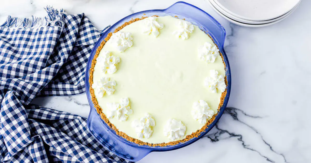 A key lime pie with a graham cracker crust and whipped cream dollops on top sits in a blue pie dish next to a blue checkered cloth and stacked white plates.