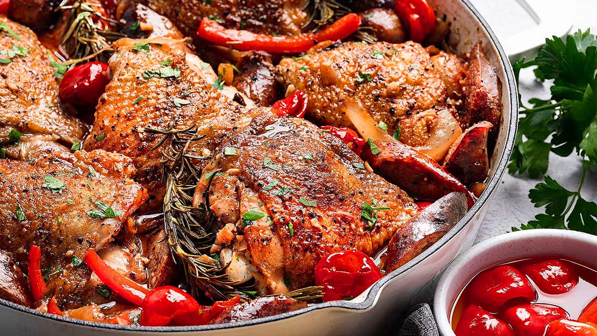 A pan filled with roasted chicken thighs, cherry tomatoes, red bell peppers, fresh herbs, and garnished with parsley sits next to a bowl of red cherry peppers.