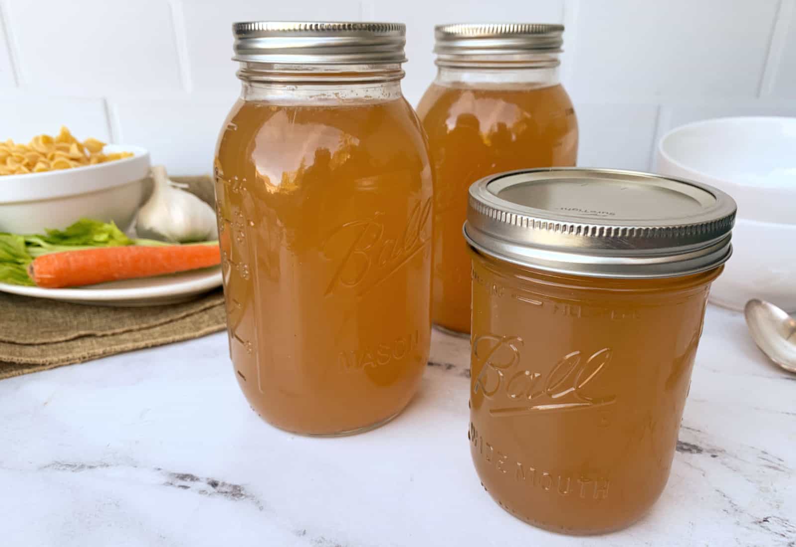 Three glass jars filled with homemade broth are sealed with metal lids and placed on a countertop, with vegetables and pasta in the background.
