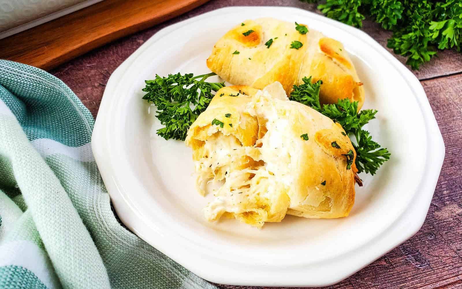 Two baked crescent rolls stuffed with creamy filling, garnished with parsley, served on a white plate with a green and white cloth beside it.