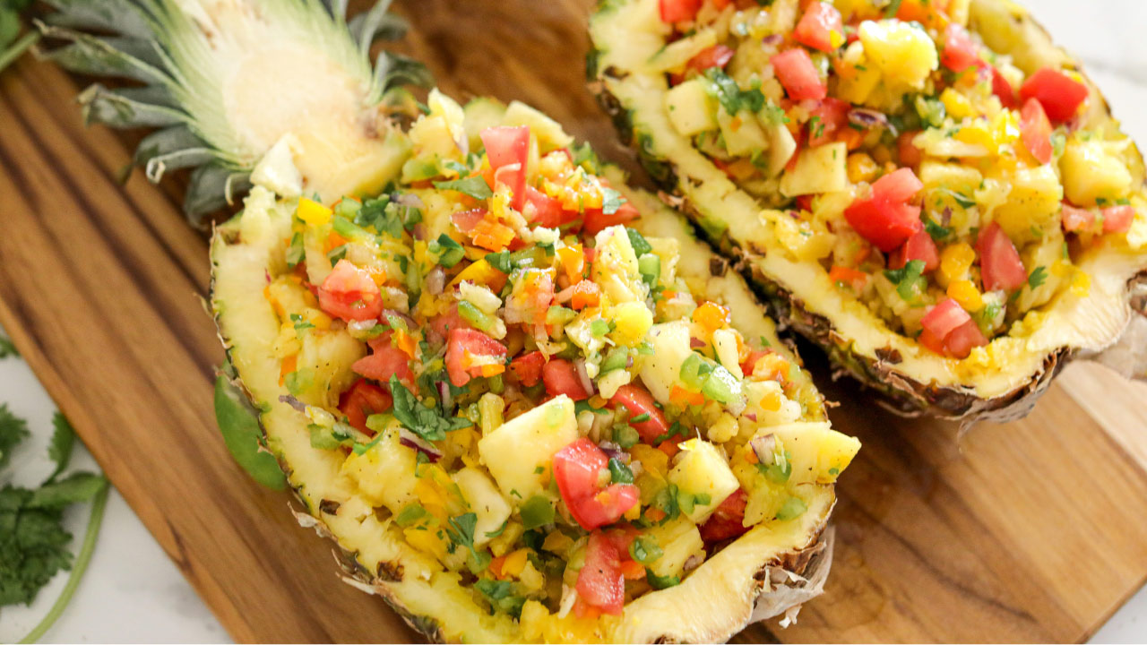 Two pineapple halves filled with a colorful mixture of diced pineapple, tomatoes, herbs, and vegetables, served on a wooden board.