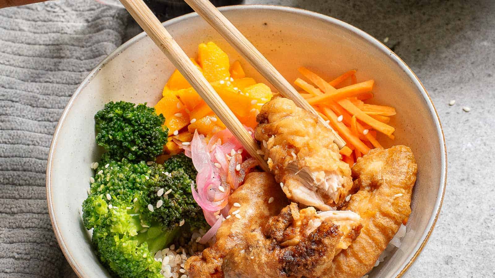 A bowl of rice topped with fried fish, broccoli, pickled onions, sliced carrots, mango, and sprinkled sesame seeds, with chopsticks holding a piece of fish.