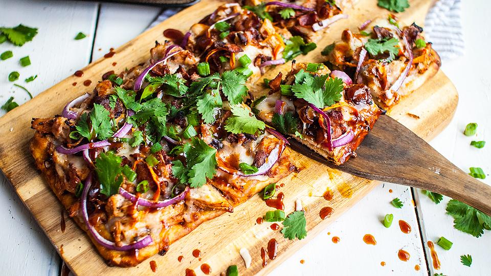 Rectangular barbecue chicken pizza topped with red onions, melted cheese, and fresh cilantro on a wooden board, with one slice being served.