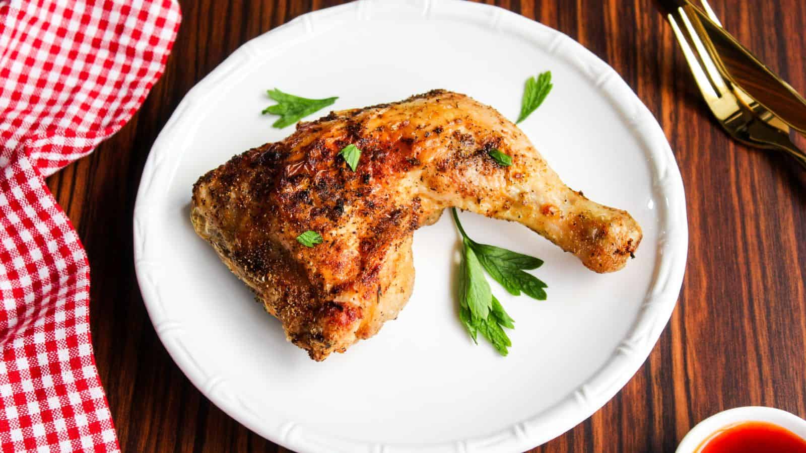 A cooked chicken leg quarter on a white plate, garnished with parsley, next to a red checkered napkin and gold utensils on a wooden table.