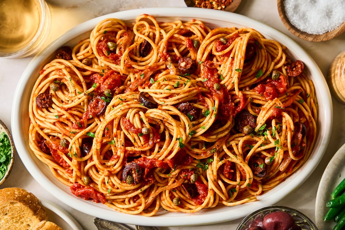A white oval dish filled with spaghetti topped with tomato sauce, olives, and herbs, surrounded by side dishes and a glass of white wine.