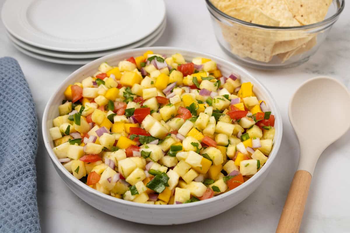 A bowl of pineapple salsa with diced pineapple, mango, red onion, tomato, and cilantro sits on a white surface next to tortilla chips, plates, a spoon, and a blue cloth.