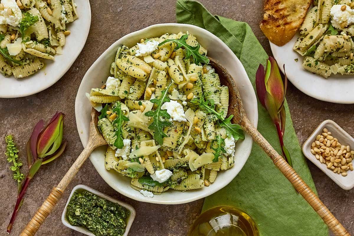 A bowl of pesto pasta topped with arugula, ricotta, and pine nuts, surrounded by plates of pasta, bread, pesto, and oil on a brown table.
