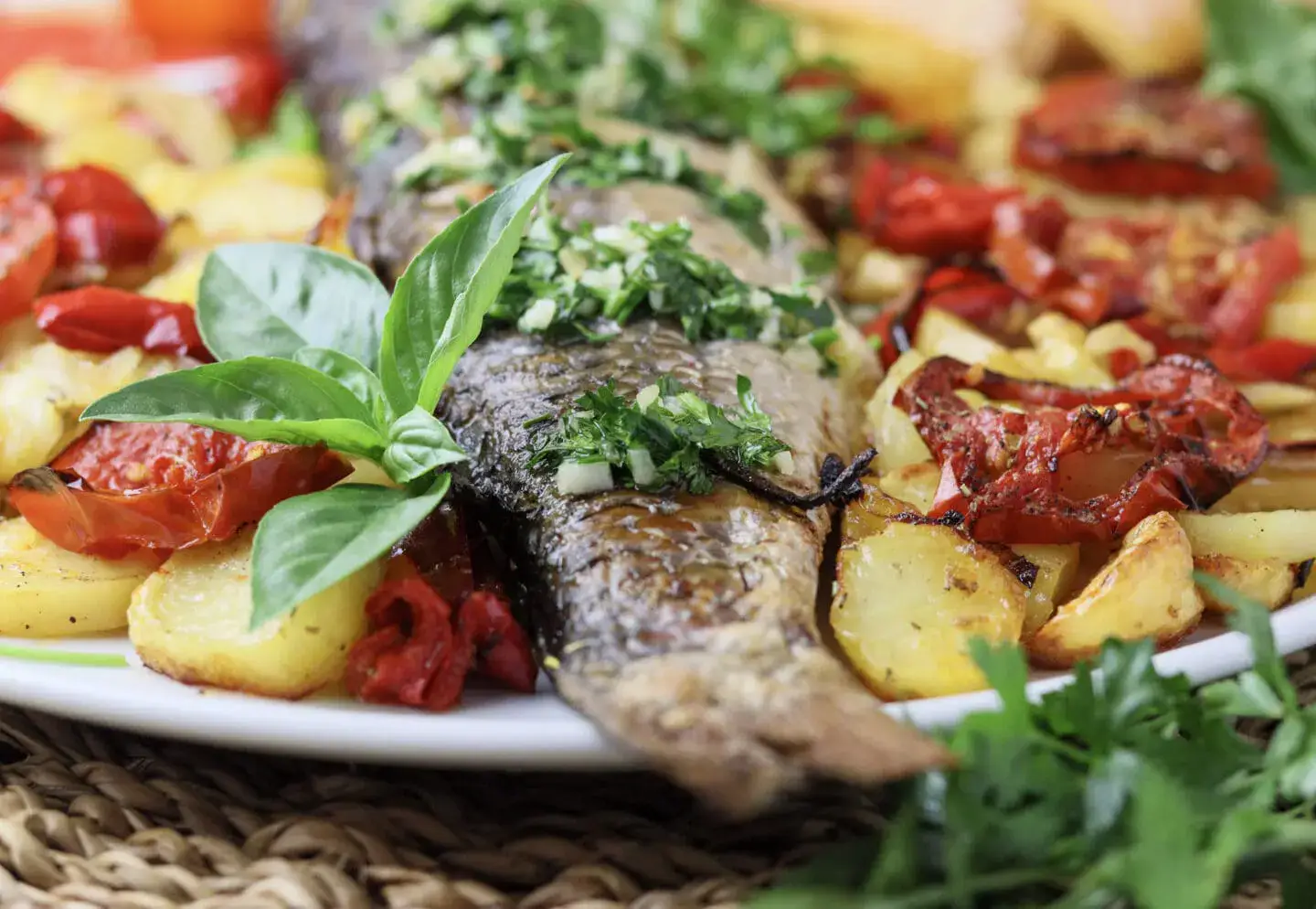A whole roasted fish garnished with chopped herbs, served with roasted potatoes, tomatoes, and fresh basil leaves on a white plate.