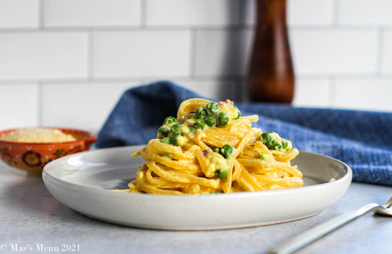 A plate of fettuccine pasta with a creamy sauce and green peas, garnished with bacon bits, sits on a white plate; a fork and a bowl of grated cheese are nearby.