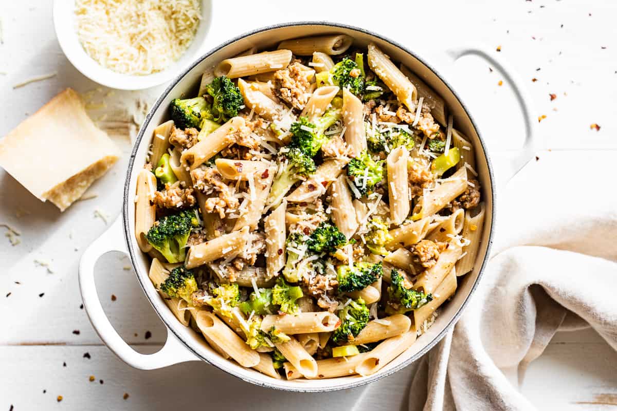 A pot of penne pasta mixed with broccoli, ground meat, and topped with grated cheese sits on a white table beside a bowl of grated cheese and a napkin.