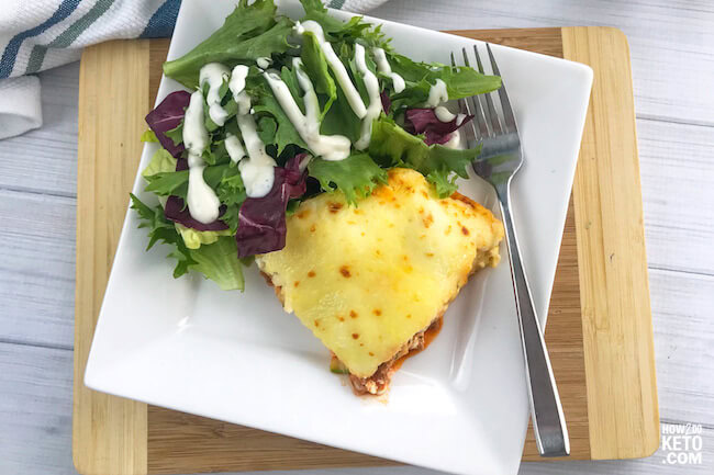 A square slice of cheesy casserole and a green salad with ranch dressing on a white plate with a fork, placed on a wooden board.