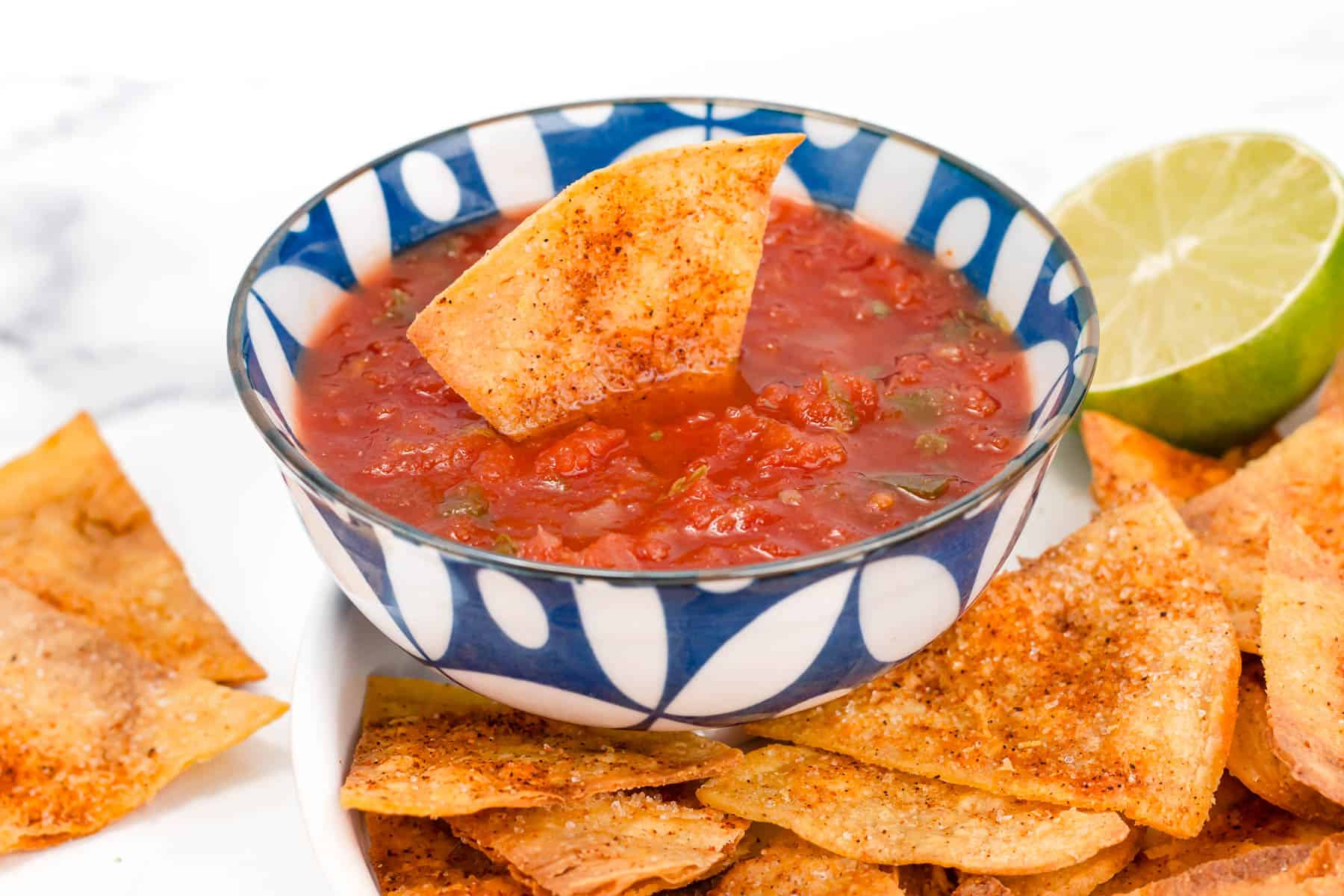 A bowl of tomato salsa with a tortilla chip dipped in it, surrounded by seasoned tortilla chips and a halved lime.