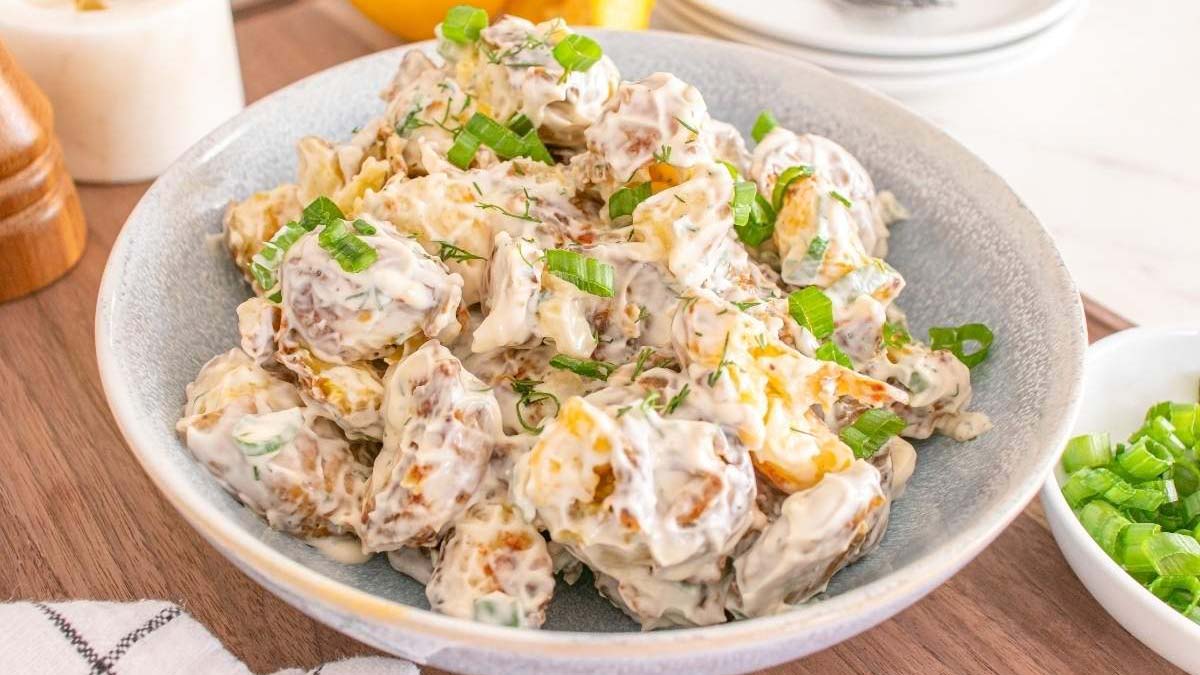 A bowl of potato salad with chopped green onions on top, mixed with a creamy dressing, sits on a wooden surface near a bowl of extra chopped green onions.