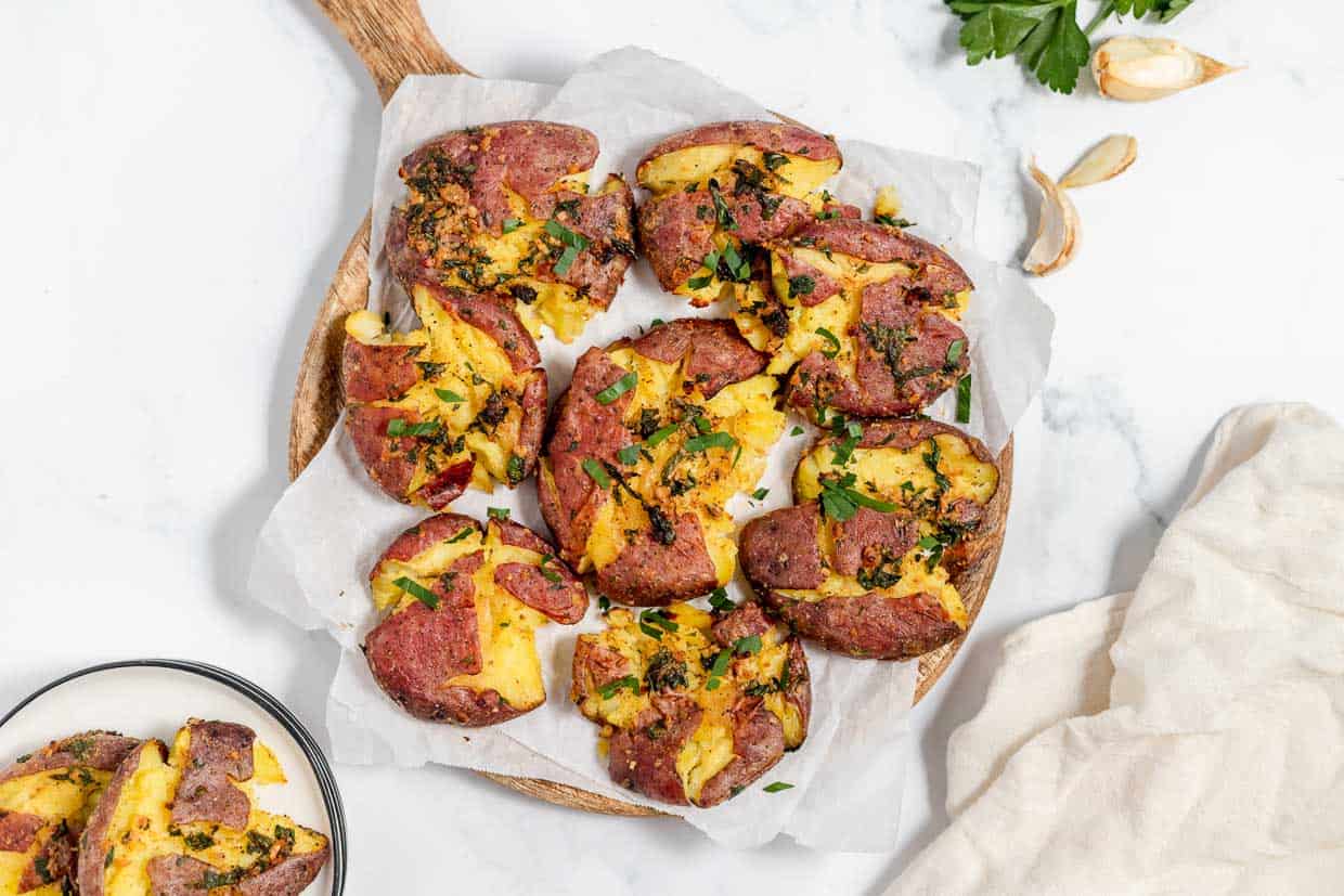 A wooden board with smashed roasted potatoes topped with herbs, displayed on parchment paper with garlic cloves and parsley nearby.