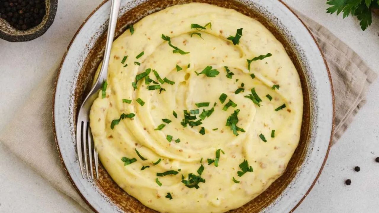 A bowl of creamy mashed potatoes garnished with chopped parsley, with a fork resting on the side of the dish.