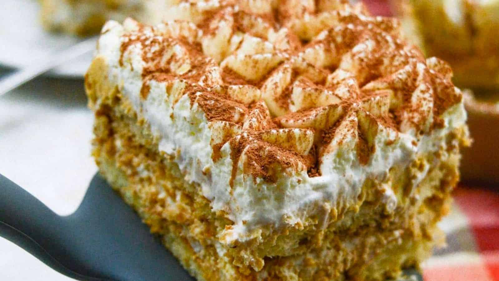 A close-up of a square slice of layered dessert with whipped cream and cocoa powder on top, served on a spatula.