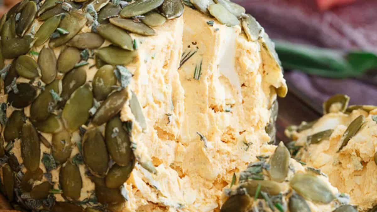 A close-up of a cheese ball coated with pumpkin seeds and herbs, showing a portion scooped out to reveal the creamy interior.