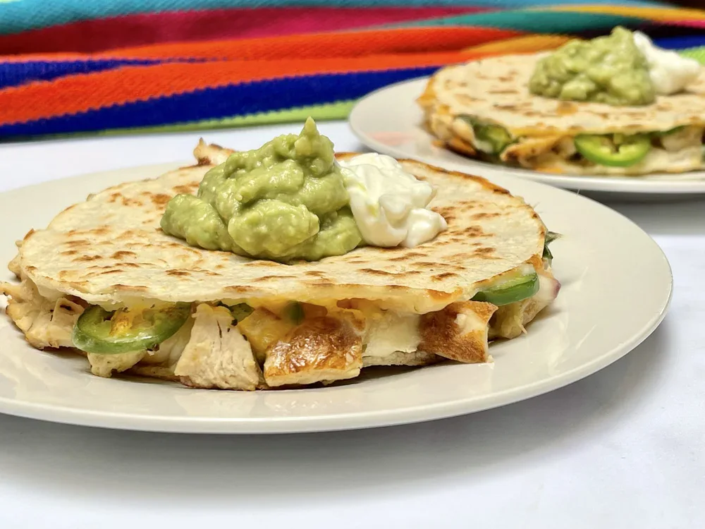 Two quesadillas filled with chicken and vegetables, topped with guacamole and sour cream, served on white plates with a colorful fabric in the background.