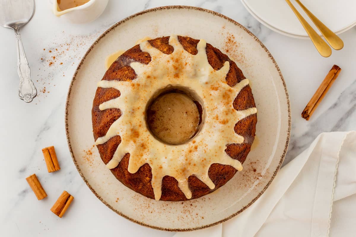 A round bundt cake with white icing drizzled on top sits on a plate, surrounded by cinnamon sticks, a spoon, and plates with utensils.