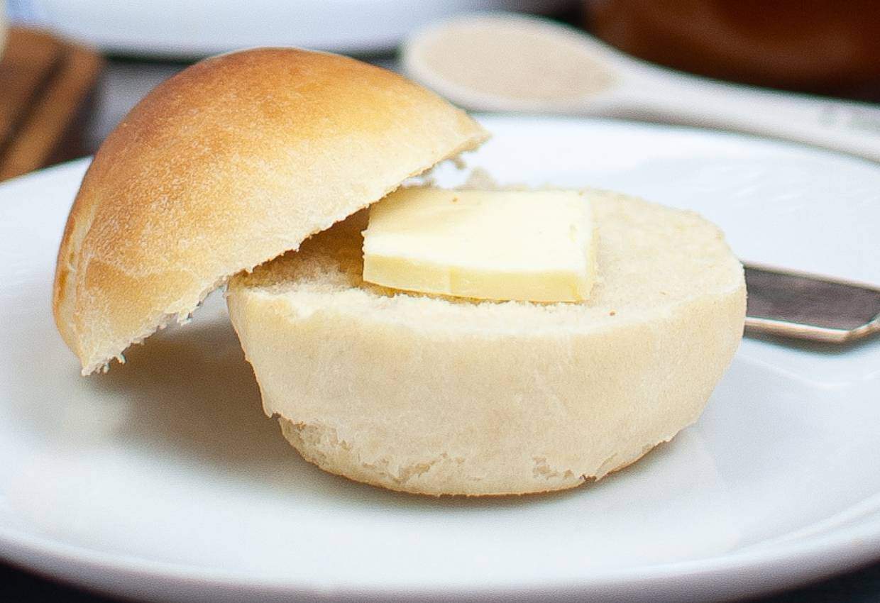 A bread roll sliced open with a pat of butter inside, placed on a white plate with a butter knife.