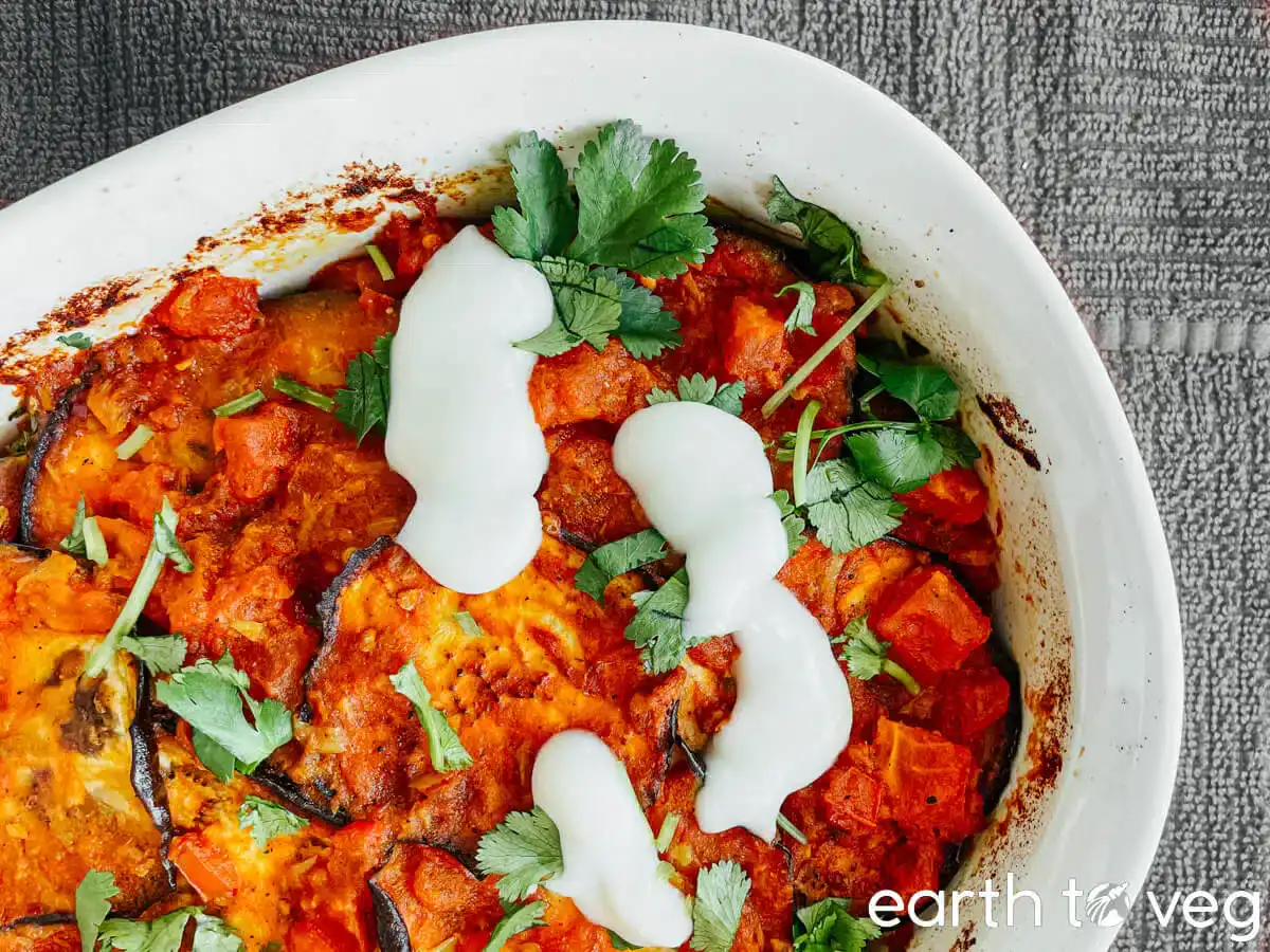 Baked vegetable casserole topped with tomato sauce, fresh cilantro, and white yogurt or cream, served in a white dish on a gray textured cloth.