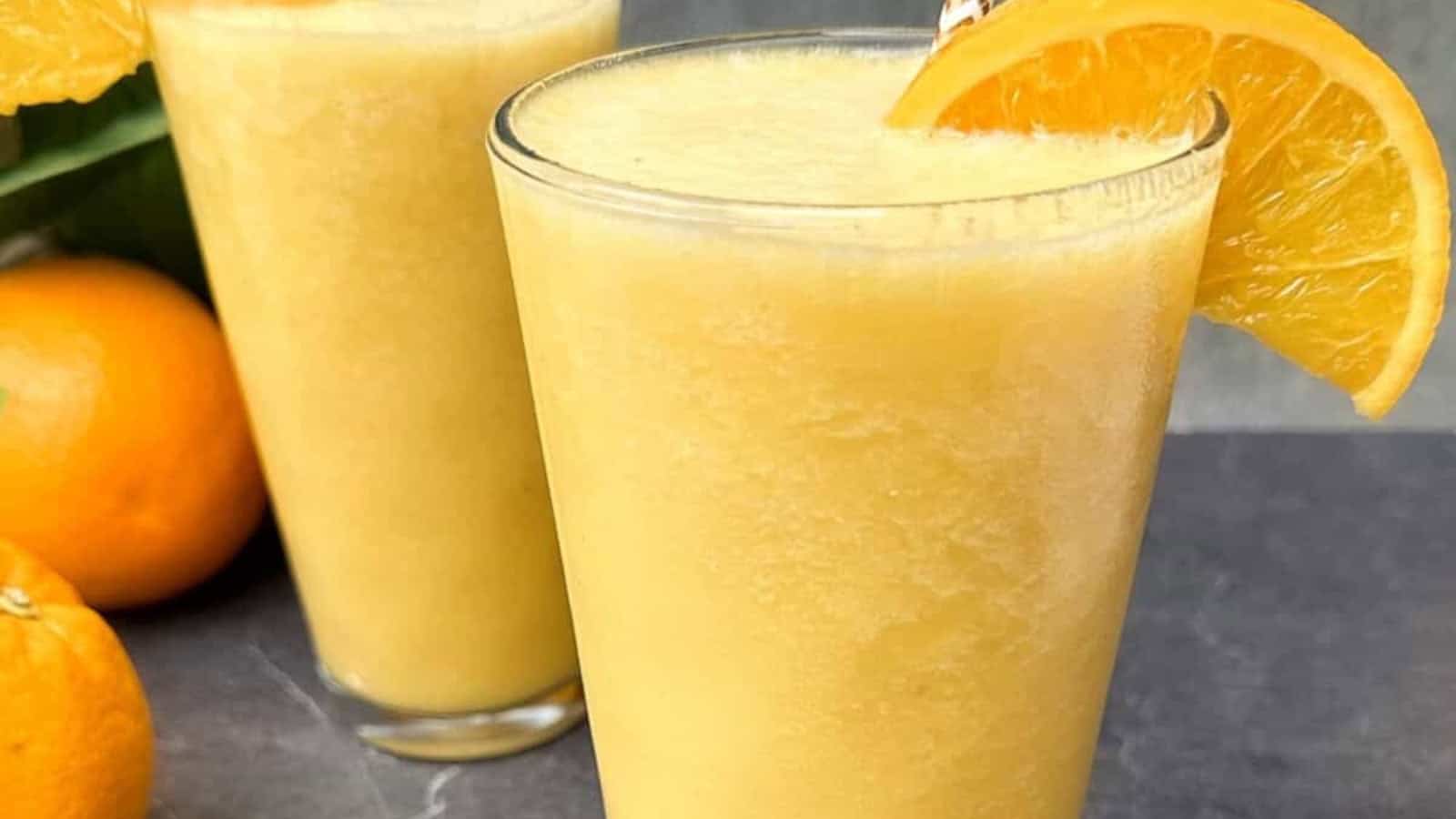 Two glasses of orange smoothie, each garnished with an orange slice, are placed on a dark surface next to whole oranges.