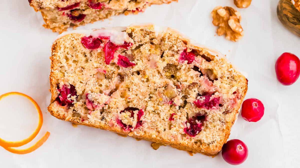 A slice of cranberry walnut bread with visible cranberries and walnuts, placed on a white surface next to whole cranberries, orange peel, and walnuts.