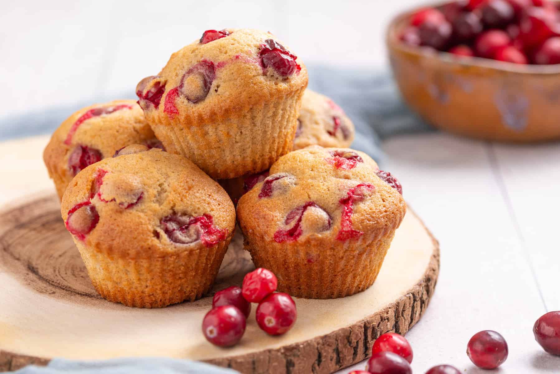 Five cranberry muffins are stacked on a wooden board, with loose cranberries scattered nearby and a bowl of cranberries in the background.