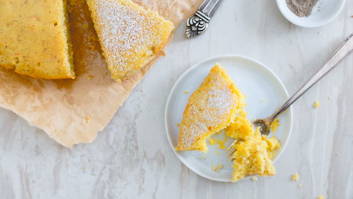 A slice of yellow cake with powdered sugar on a small plate and a fork, next to the rest of the cake on parchment paper.