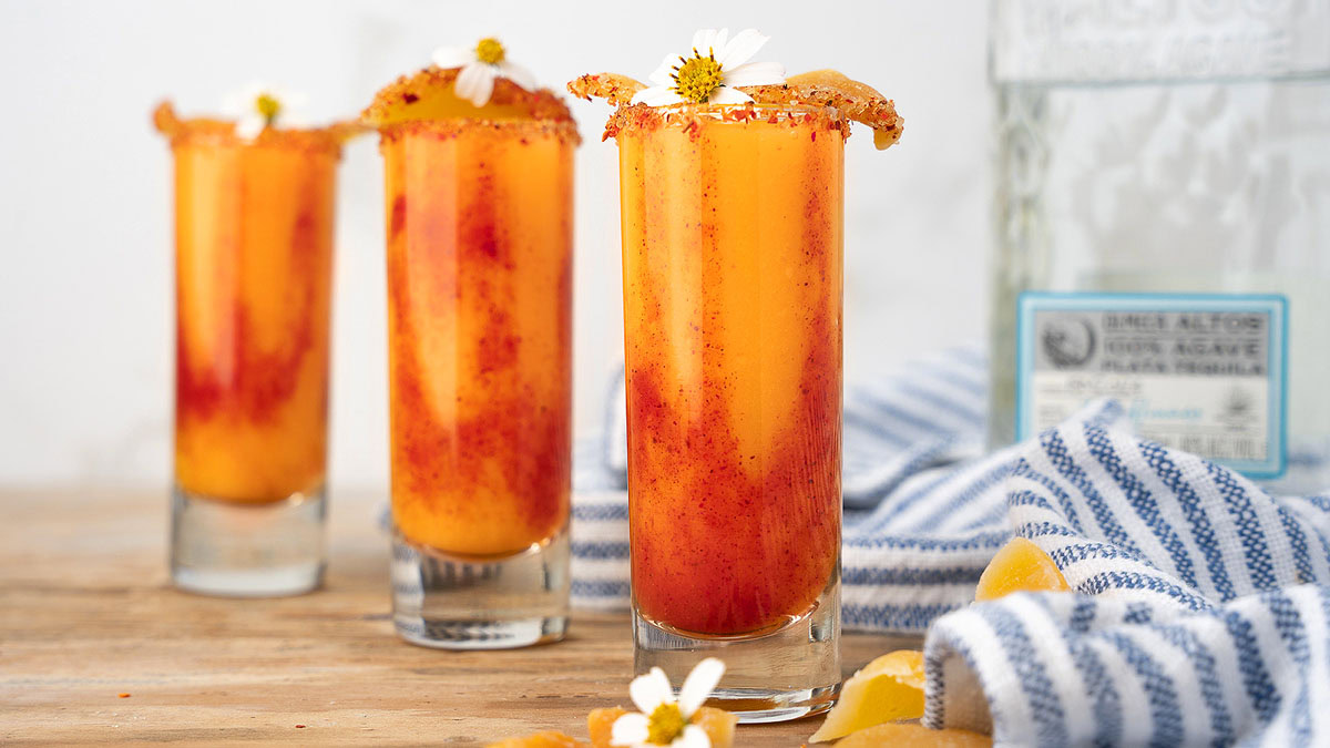 Three tall glasses filled with an orange and red layered drink, garnished with flowers and chili powder on the rim, with a tequila bottle and striped towel in the background.