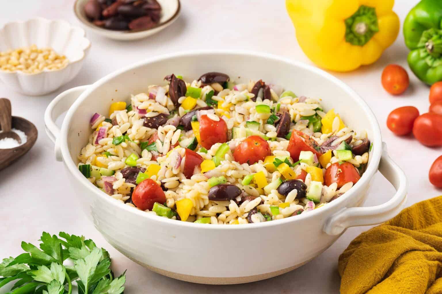 A bowl of orzo pasta salad with cherry tomatoes, olives, diced cucumbers, yellow bell peppers, and red onions, surrounded by fresh vegetables and herbs on a table.