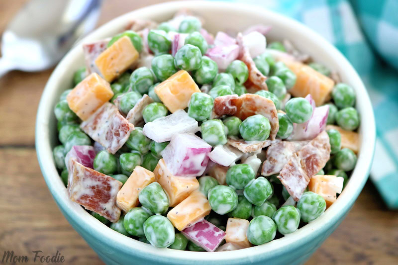 A bowl of salad with green peas, cheddar cheese cubes, diced red onions, and bacon pieces, coated in a creamy dressing.