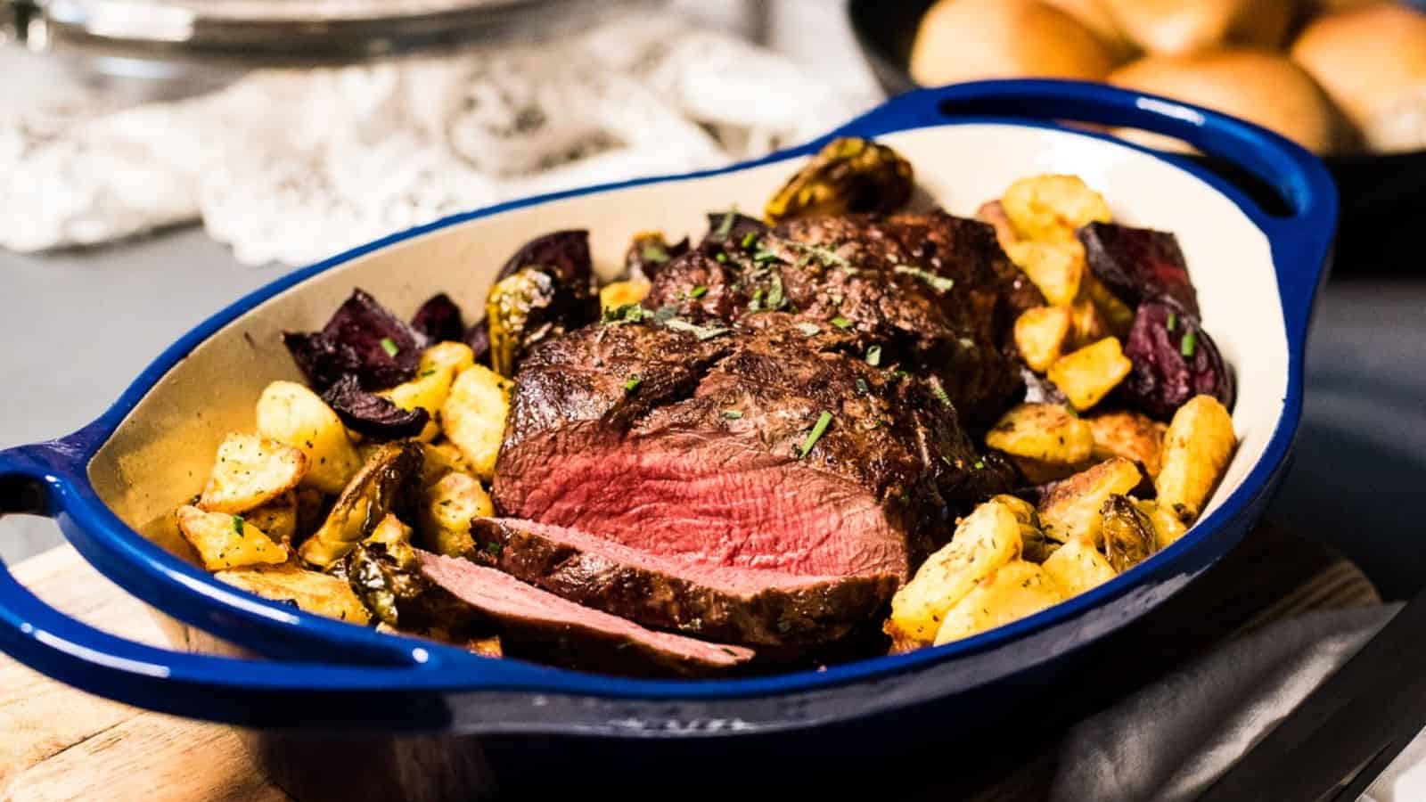 Sliced medium-rare roast beef served in a blue dish with roasted potatoes and vegetables.