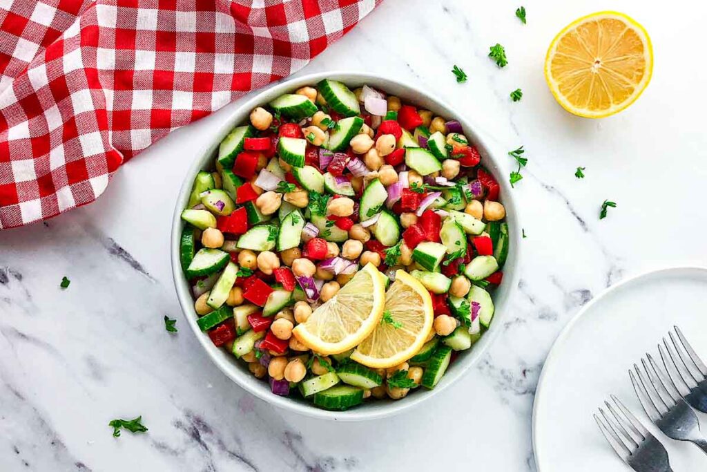 A bowl of chickpea salad with cucumbers, red peppers, onions, and lemon slices, placed on a marble surface next to a halved lemon, forks, and a red checkered cloth.