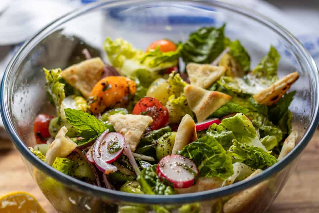 A glass bowl filled with fattoush salad, featuring lettuce, cherry tomatoes, radishes, fresh herbs, and toasted pita chips.