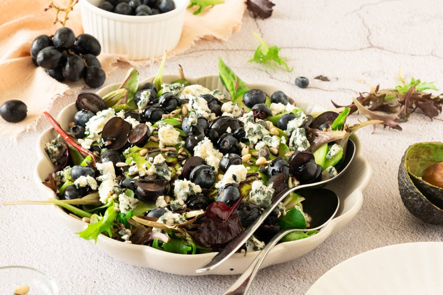 A salad with mixed greens, sliced black grapes, crumbled blue cheese, and seeds is served in a bowl with a spoon and fork. Blueberries and grapes are on the table nearby.