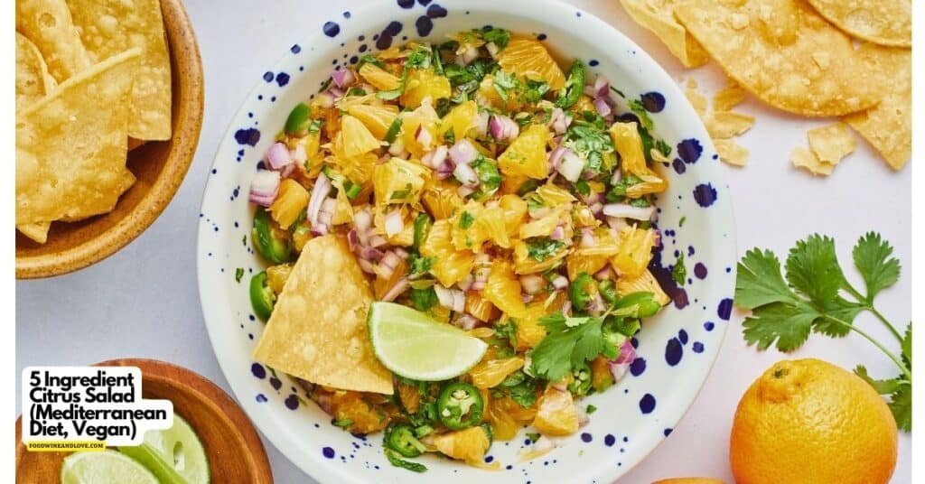 A bowl of citrus salad with chopped oranges, red onion, cilantro, and jalapeños, served with tortilla chips and a lime wedge on the side.