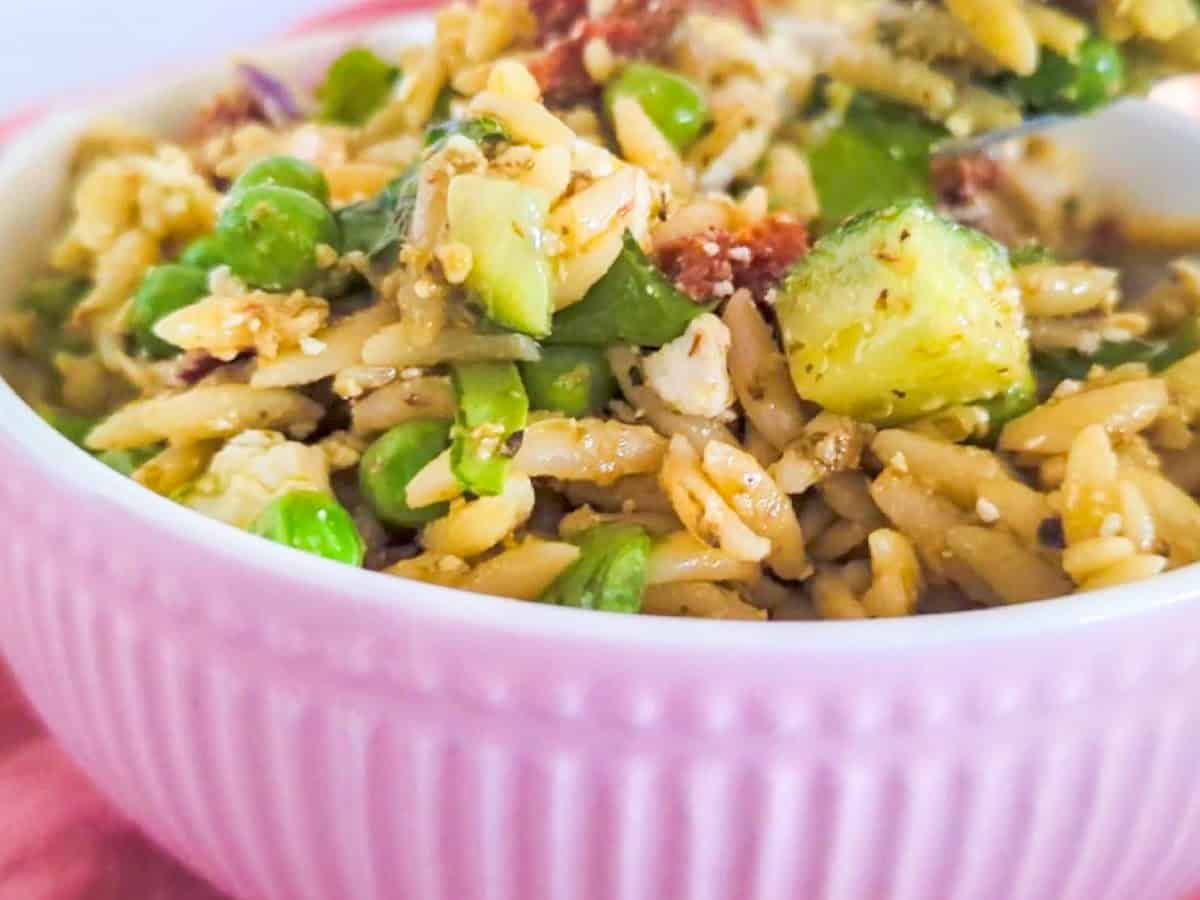 A close-up of a bowl filled with orzo pasta, peas, zucchini, and crumbled cheese in a light pink bowl.