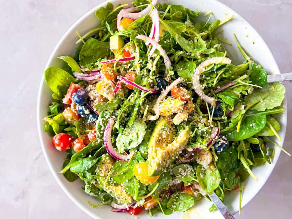 A bowl of mixed green salad with spinach, cherry tomatoes, avocado, red onion, blueberries, and sesame seeds, served with a fork.